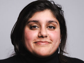 Shama Ali wearing a black turtleneck, standing against a plain gray background.