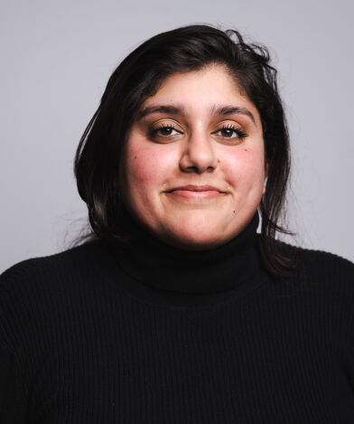 Shama Ali wearing a black turtleneck, standing against a plain gray background.